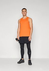 Narancssárga trikó, fekete rövidnadrág, fekete leggings és fekete sportcipő. A trikón logó található; a leggings diszkrét fényes felületű. Edzőterem környezet.