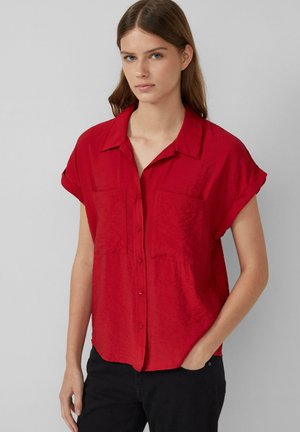 Camicetta rossa a maniche corte con colletto, dotata di due tasche sul petto e realizzata in un tessuto leggero e testurizzato. Indossata con pantaloni scuri.