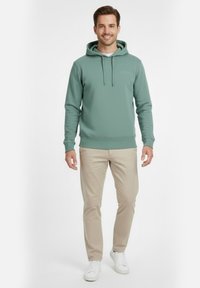 Sudadera con capucha verde claro con acabado texturizado, cordón ajustable y bolsillo frontal tipo canguro, combinada con pantalones beige y zapatillas blancas.