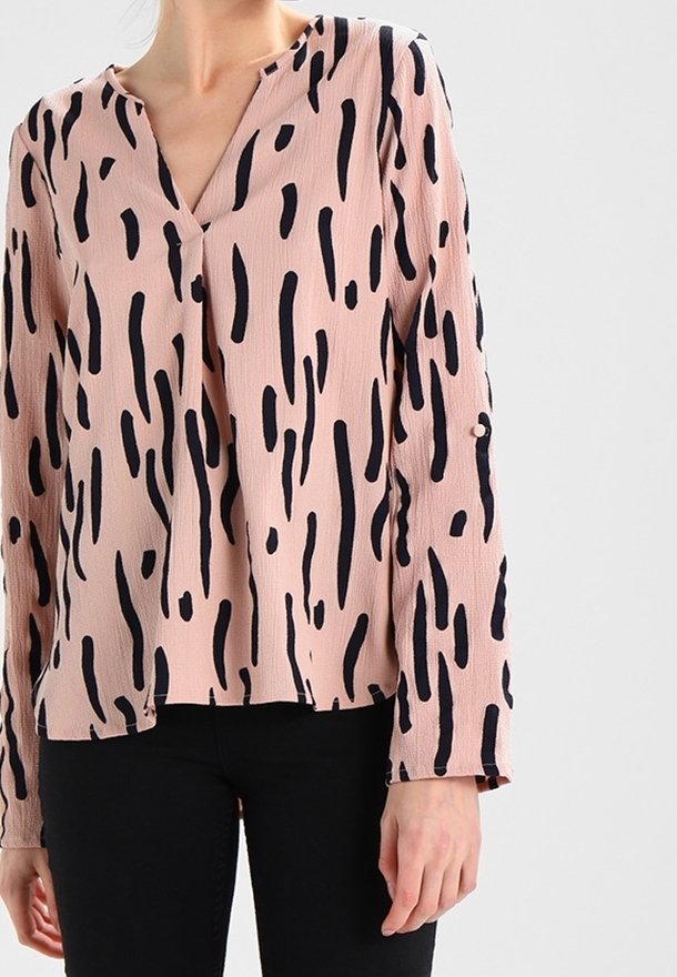 Femme portant un chemisier rose clair avec des rayures verticales noires irrégulières et un pantalon noir, visible du cou aux hanches sur un fond uni.