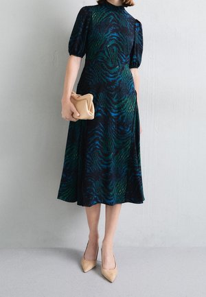 Femme portant une robe midi à motifs bleu foncé et vert avec des manches bouffantes, tenant une pochette beige et portant des escarpins beiges à bouts pointus.