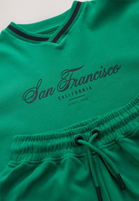 Green sweatshirt with a v-neck, black trim, and "San Francisco" print. Paired with matching shorts featuring an elastic waistband and drawstring.