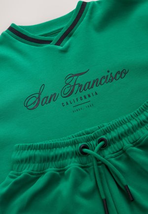 Grøn sweatshirt med V-hals, sort kant og "San Francisco"-tryk. Kombineret med matchende shorts med elastisk linning og snøre.