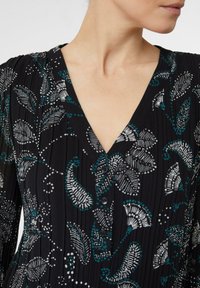 Frau trägt eine schwarze V-Ausschnitt-Bluse mit weißen und türkisfarbenen Blumen- und Blattmustern sowie vertikalen Falten.