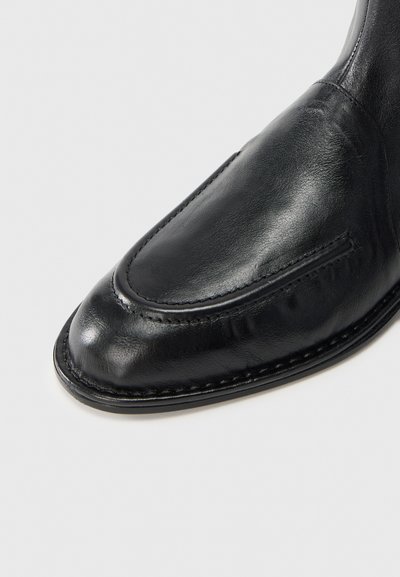 Μαύρο δερμάτινο παπούτσι με στρογγυλεμένη μύτη, λεία επιφάνεια, διακοσμητικές ραφές και ελαφρώς ανυψωμένη φτέρνα.
