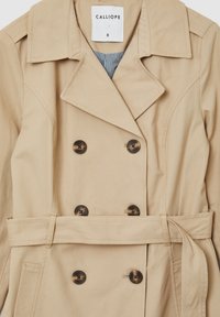 Beige Trenchcoat mit doppelter Knopfleiste, großen Revers und einem Taillengürtel. Mit gestreiftem Innenfutter und zwei Seitentaschen.