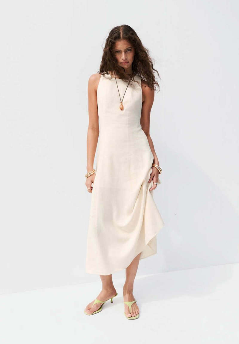 Femme aux longs cheveux bouclés portant une robe midi crème sans manches, des sandales vertes à talons, un collier pendentif et des bracelets en or aux deux poignets.