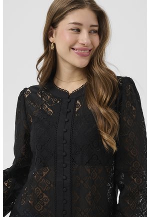 Jauna sieviete ar gariem viļņotiem matiem, kas smaida, valkājot zelta riņķa auskarus un melnu mežģīņu blūzi ar pogām un ziedu rakstiem.