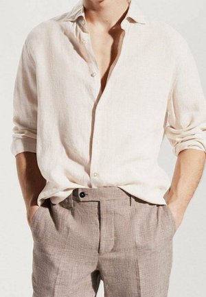 Hombre vistiendo una camisa de lino beige clara con las mangas remangadas y pantalones de vestir a cuadros beige, con las manos casualmente en los bolsillos.