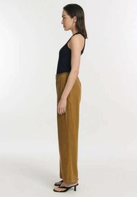 Zwarte mouwloze top, bruine hoge taille broek, slanke pasvorm, licht gestructureerde stof en dunne zwarte haksandalen. Zijdelings profielzicht.