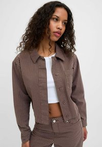 Brown striped jacket with a button-up front and collar, featuring a chest pocket. Worn over a white crop top, with long sleeves.