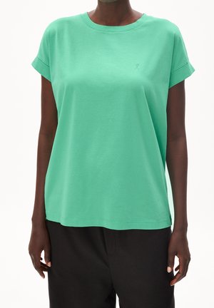 Nainen pukeutuneena lyhythihaiseen mintunvihreään puseroon, jossa on pyöreä pääntie, ja mustiin housuihin, valkoista taustaa vasten.