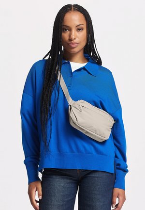 Donna con lunghe trecce che indossa un maglione blu, jeans scuri e una borsa beige a tracolla su uno sfondo bianco semplice.