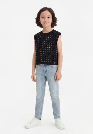 Vaikas su pečius siekiančiais plaukais stovi dėvėdamas juodą be rankovių viršutinį rūbą su mažais baltais kvadratais, šviesiai mėlynas džinsus ir baltus sportinius batelius.