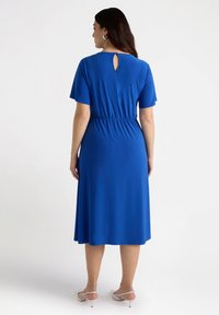 Sininen midi-mekko, jossa on lyhyet hihat, elastinen vyötärö ja avokolo takana. Sujuva kangas ja virtaava siluetti.