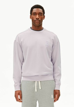 Mann steht, trägt ein helllila Sweatshirt mit Rundhalsausschnitt und graue Jogginghose mit weißen Kordeln, vor einem schlichten weißen Hintergrund.