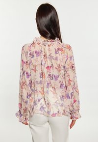Blumige Bluse mit einem durchsichtigen, leichten Stoff und Rüschenausschnitt sowie -ärmeln. Bunte lila und pinke Blumenmuster auf einem hellen Hintergrund.
