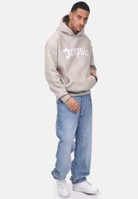 Beige Kapuzenpullover mit weißem "Dropsia"-Logo. Getragen mit hellblauen Jeans und weißen Sneakern. Verfügt über eine Vorderseite Tasche und einen lockeren Schnitt.