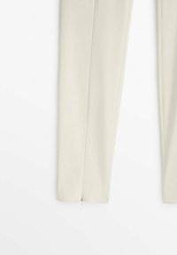 Gros plan sur les jambes d’un pantalon beige clair avec des coutures latérales visibles et de petites fermetures éclair argentées aux ourlets inférieurs sur fond blanc.