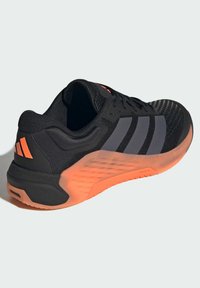 Svart treningssko med oransje detaljer, en pustende mesh-overdel og en teksturert gummisåle. Har tre grå striper og en polstret hæl.