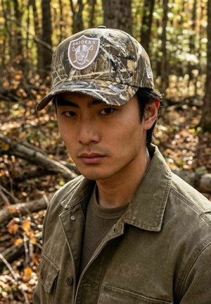 Young man in a camo Raiders cap and olive jacket standing in a wooded area with fallen leaves and trees in autumn light.