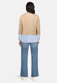 Veste en daim beige à manches trois-quarts, associée à une chemise rayée bleu et blanc, un jean évasé bleu clair et des baskets blanches.