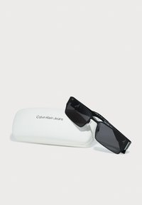 Juodos saulės akinių su stačiakampiais rėmeliais ir tamsiais lęšiais, kurie kartu su balta kietąja dėžute, ant kurios juodu šriftu parašyta "Calvin Klein Jeans".