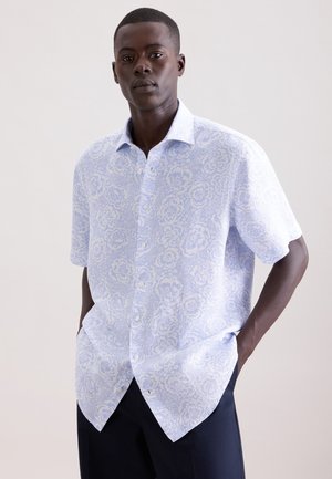 Man draagt een lichtblauw met wit bloemenpatroon overhemd met korte mouwen, met handen in de zakken van een marineblauwe broek, tegen een effen achtergrond.