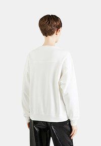 Felpa bianca con spalle scese e collo a giro. Presenta una texture liscia e una vestibilità rilassata. Indossata con pantaloni neri lucidi e stampati.