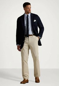 Mężczyzna w granatowej marynarce, niebieskiej koszuli w kratę, granatowym krawacie, beżowych spodniach i brązowych mokasynach, stoi z jedną ręką w kieszeni.