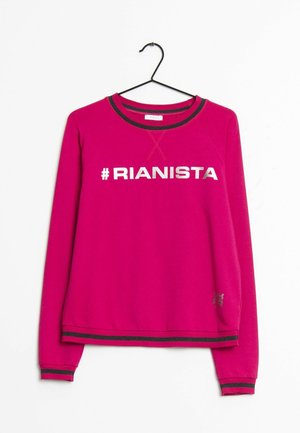 Jasnoróżowa bluza z długim rękawem z czarną lamówką, z białym napisem "#RIANISTA" na piersi, zawieszona na czarnym wieszaku.