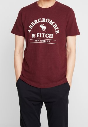 Osoba ubrana w bordową koszulkę Abercrombie & Fitch z białym logo i czarne spodnie, ręce w kieszeniach, na jednolitym tle.