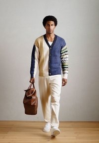 Strikket cardigan med en blanding af blå, beige og mønstrede ærmer; parret med hvide bukser og bærende en brun læder sportstaske.