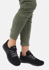 Baskets en feutre noir avec des lacets gris, forme de bout arrondi et semelle en caoutchouc flexible. Pantalon vert olive, à coupe fuselée et ourlet à revers.