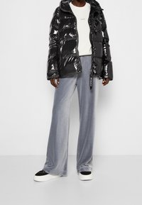 Černá lesklá prošívaná bunda s kapucí, oblečená přes světle šedou mikinu a šedé sametové kalhoty s širok ými nohavicemi, doplněná černo-bílými teniskami.