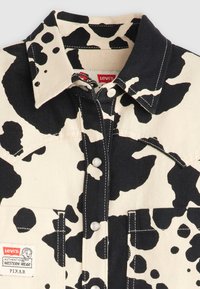 A Levi's tehénmintás ing bézs alapszínnel és fekete foltokkal rendelkezik, farmer anyagból készült, gombos elülső résszel és egy mellzsebbel, amelyen egy márkajelzés található.