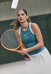 Teal sporttop van lichtgewicht stof, met een wit Nike-logo, gecombineerd met een witte geplooide tennisskirt. Oranje racket in de handen.