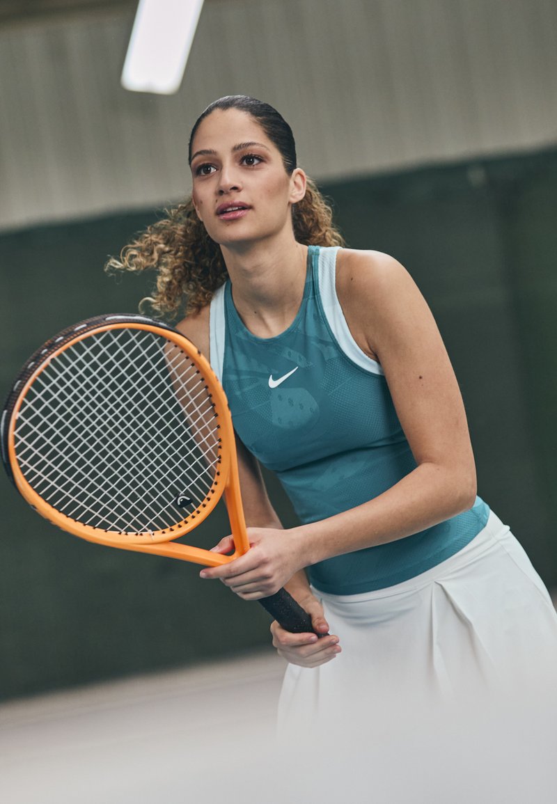 Teal sporttop van lichtgewicht stof, met een wit Nike-logo, gecombineerd met een witte geplooide tennisskirt. Oranje racket in de handen.