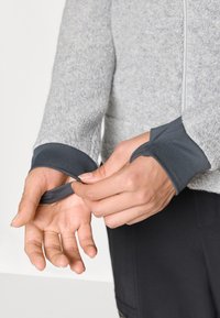 Veste en polaire grise avec des poignets à trou pour le pouce gris foncé. Des mains ajustent le poignet, mettant en valeur la douceur du tissu et le design sportif. Fermeture éclair à l'avant visible.