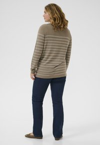 Femme aux cheveux ondulés mi-longs portant un pull rayé taupe, un jean bleu foncé et des chaussures plates marron, se tenant de profil vers la gauche.