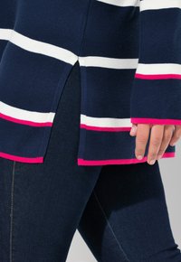 Sweter w marynarski paski z białymi i różowymi akcentami, z rozcięciem na dole i prążkowaną teksturą. Połączony z ciemnogranatowymi dżinsami.