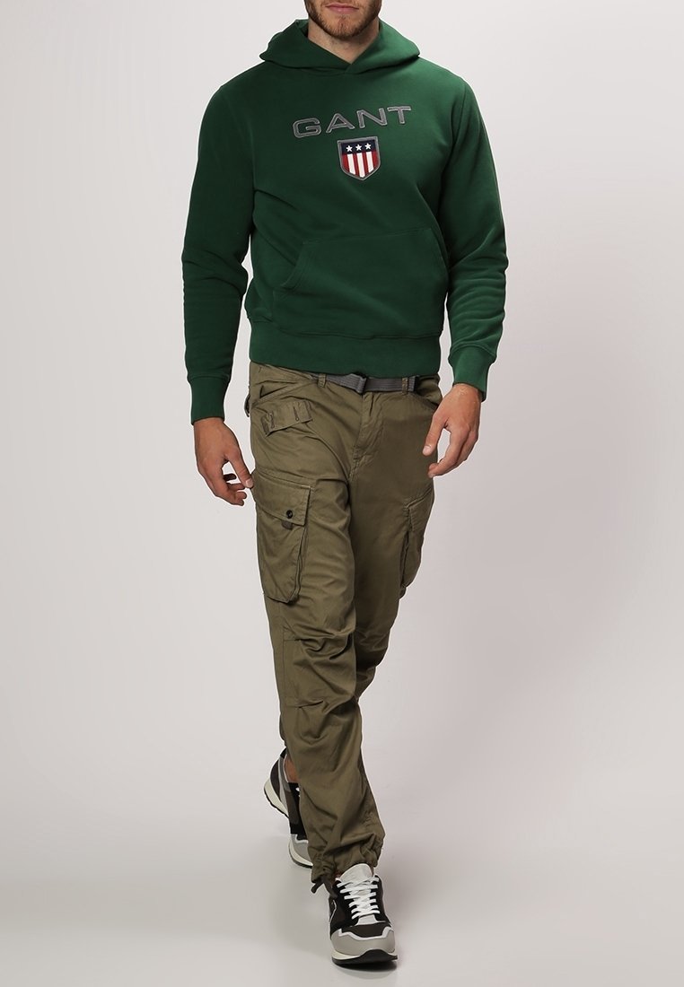 Homme marchant portant un sweat à capuche vert GANT, un pantalon cargo olive, des baskets noires et blanches, sur un fond clair uni.
