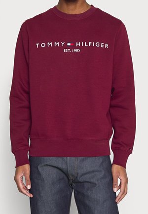 Osoba ubrana w bordową bluzę z napisem "Tommy Hilfiger EST. 1985" i logo haftowanym na piersi, w połączeniu z ciemnoniebieskimi dżinsami.