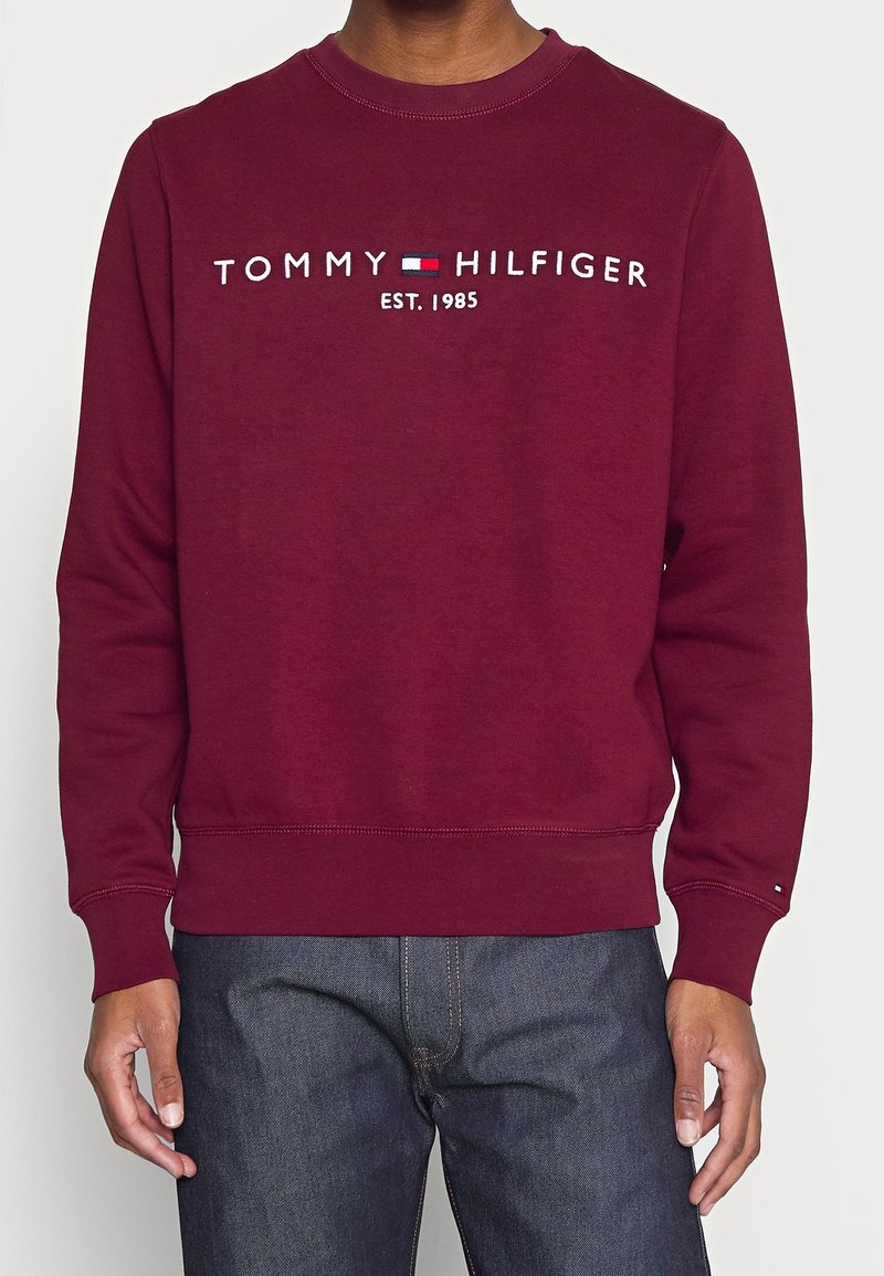 Personne portant un sweat-shirt bordeaux avec "Tommy Hilfiger EST. 1985" et le logo brodés sur la poitrine, associé à un jean bleu foncé.