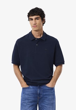 Jonge man in een donkerblauw polo-shirt en blauwe jeans, staand met de handen in zijn zakken tegen een witte achtergrond.
