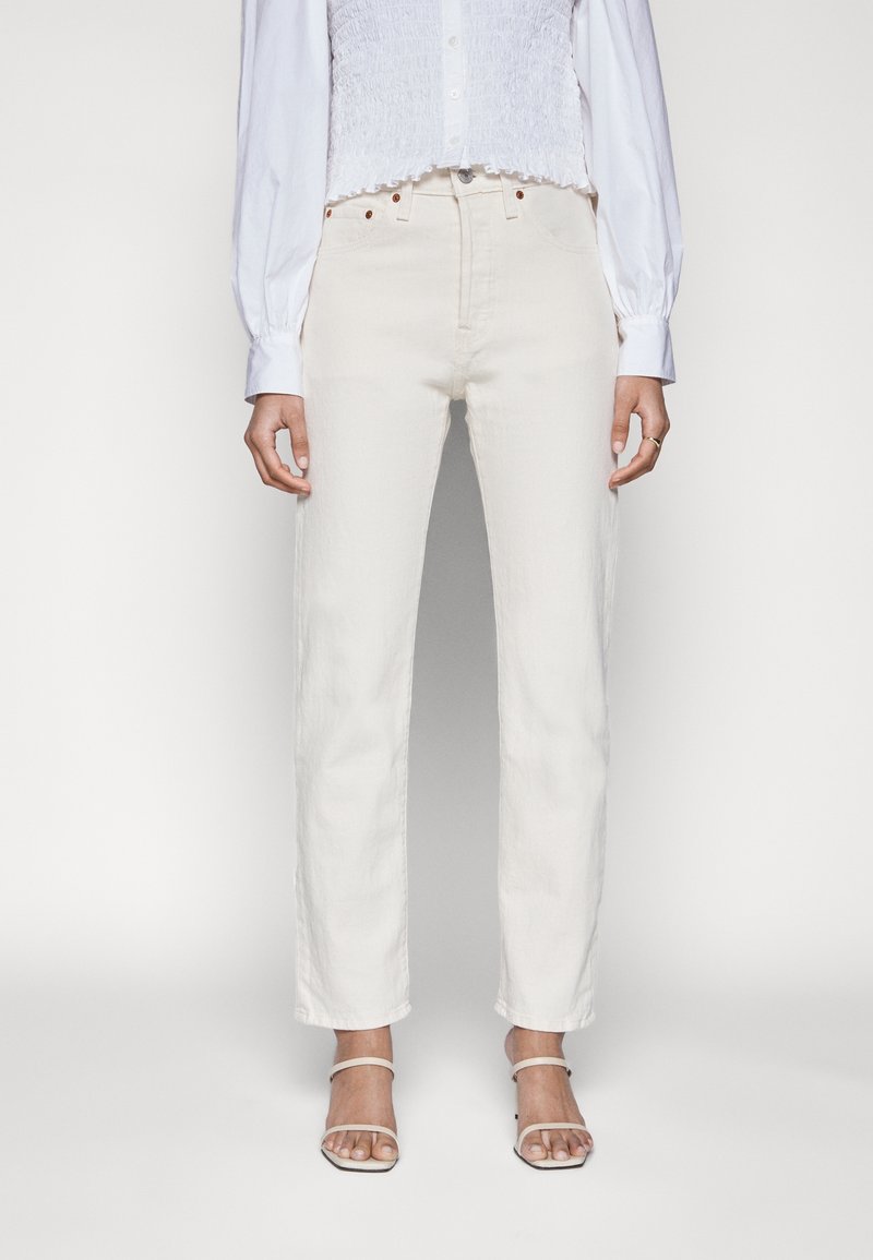 Femme portant un jean blanc droit taille haute, un chemisier blanc boutonné à manches longues et des sandales blanches à talons avec des brides.