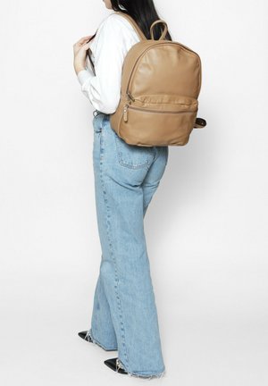 Sac à dos en cuir tan avec une forme arrondie, une poignée supérieure et des bretelles ajustables ; il dispose d'une poche zippée à l'avant et d'une texture lisse.