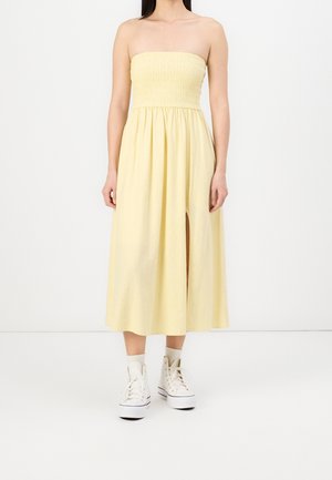 Mujer con vestido midi amarillo sin tirantes con abertura frontal y zapatillas blancas de caña alta sobre fondo blanco liso.