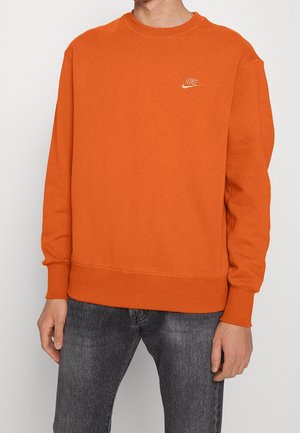Mand iført orange Nike-sweatshirt med lille logo på brystet og mørkegrå jeans, stående mod en ensartet lys baggrund.