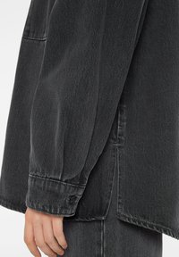 Black denim jacket with a relaxed fit, featuring a button cuff, visible stitching, and a textured fabric, side slit at the hem.